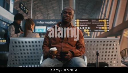Black Man Sits in Airport Lounge, Using Smartphone Scan QR Code on Ticket. While Waiting Flight, Male Traveler Drinks Cup of Coffee, Calmly Double Checks Departure Details. Modern Travel Technology. Stock Photo
