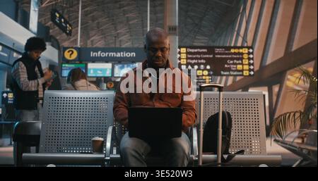African American Man Sits in Terminal Hall, Concentrated on Laptop Screen, Waiting For Departure, With Bags and Coffee Nearby. Digital Nomad Lifestyle, Telecommuting, Global Connectivity of Traveler. Stock Photo