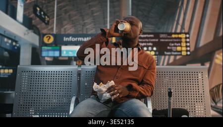 Man Takes Break From Journey Have Quick Snack. Sitting in Airport Waiting Area With Luggage Nearby, Male Unwraps Sandwich and Drinks Coffee in Paper Cup. Authentic Atmosphere of Long Transit Hours. Stock Photo