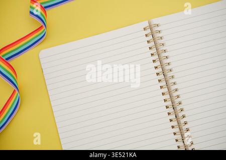 Rainbow ribbon lies next to open notebook with blank lined pages on yellow background. Education and inclusivity. Stock Photo
