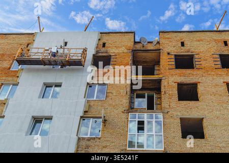 An empty construction cradle hangs on a war-damaged brick apartment building, part of the facade is newly insulated and windows are installed Stock Photo