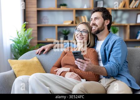 Smiling young adult couple sitting together on a comfortable sofa at home, enjoying a moment of leisure while looking at a smartphone screen and sharing a digital experience Stock Photo
