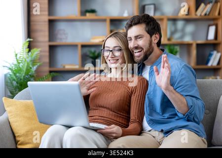 Smiling couple on a sofa waving at a laptop during a cheerful video call, sharing happy moments and staying connected with friends or family from their cozy living room Stock Photo