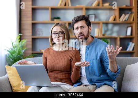 Young couple looking puzzled and shrugging while experiencing technical issues or an error on a laptop computer, sitting on a sofa in their living room Stock Photo