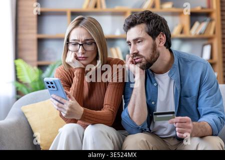 Worried couple sitting on a sofa looking at a smartphone, holding a credit card and experiencing stress about financial problems, online shopping, and accumulated debt Stock Photo