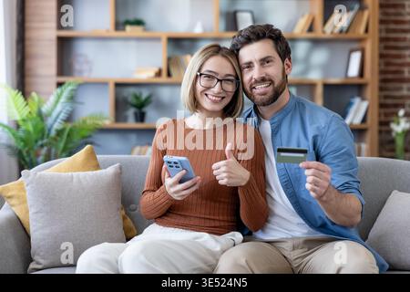 Smiling young couple relaxed on a sofa, using smartphone and credit card for easy, secure online shopping at home modern mobile banking and convenient e-commerce comfort together Stock Photo