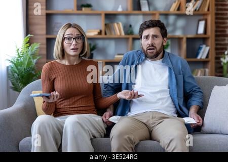 Frustrated young couple sitting on a sofa at home, facing financial difficulties and showing empty pockets, feeling stressed about bankruptcy and money problems Stock Photo