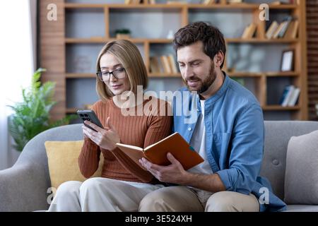 Couple sitting on a couch, collaborating on household tasks, with the woman using a smartphone and the man reading a notebook, both engaged in financial planning and online activities Stock Photo