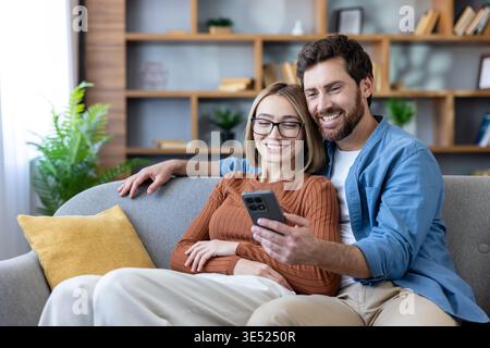 Cheerful young couple is sitting on a cozy sofa at home, scrolling through mobile applications on a smartphone and enjoying their relaxed shared time in the living room Stock Photo