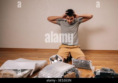 Puzzled man kneeling on hardwood floor, expressing extreme frustration and stress while attempting to assemble new office chair from complex flat pack Stock Photo