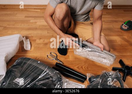 Contractor male unboxing and assembling new flat-pack office chair for home office, carefully unpackaging components on hardwood floor, engaging in Stock Photo