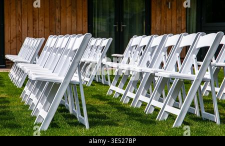Rows of white folding chairs arranged on a green lawn for an outdoor wedding ceremony in front of a modern building with wooden cladding Stock Photo