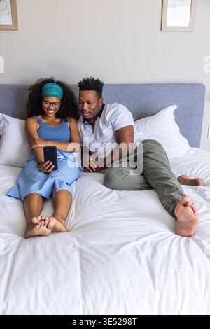 Diverse couple lounging on bed at home, woman holding smartphone wearing glasses and headwrap Stock Photo