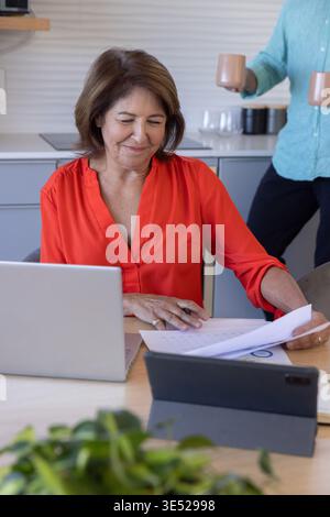 Couple in kitchen, senior red top marking papers at devices with pen, man holding mugs, plant Stock Photo