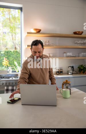 Mature adult Asian man standing at kitchen island wearing sweater reaching smartphone, using laptop Stock Photo