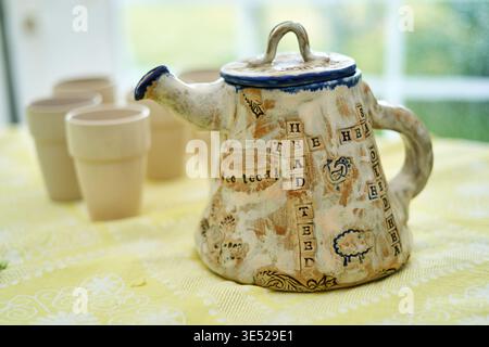 A rustic handmade ceramic teapot with a crossword puzzle design sits prominently on a yellow floral tablecloth. The teapot features a distressed Stock Photo