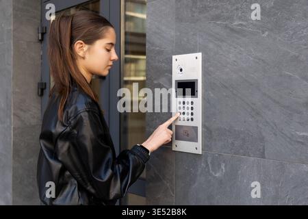Young woman using intercom at apartment entrance. Young woman presses a video intercom keypad at a modern apartment entrance, illustrating residential Stock Photo