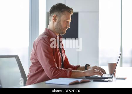 Man Working on Laptop in Bright Office With Cup of Coffee and Notebook on Table Stock Photo
