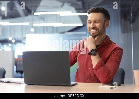 Man Sitting at Desk Working on Laptop and Smiling During Online Meeting in Office Setting Stock Photo