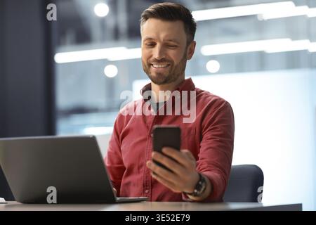 Man Using Smartphone at Desk While Working on Laptop in Modern Office Space Stock Photo