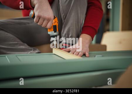 Close-up of a man's hands operating a cordless power drill to screw a wooden piece into a new piece of furniture, representing diy home improvement an Stock Photo