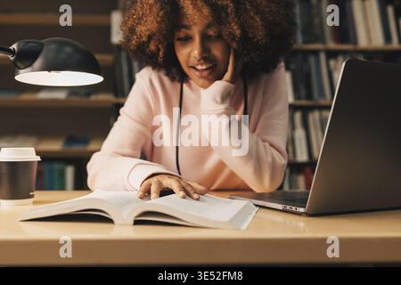 Smiling girl preparing exams in library at evening. Student sitting at desk and reading Stock Photo