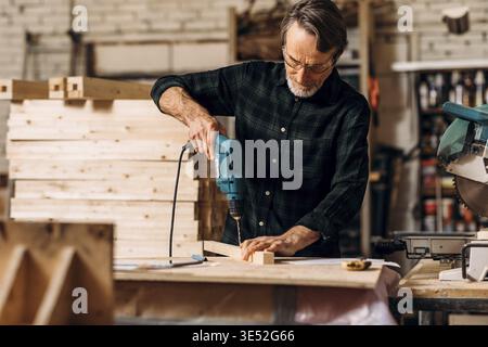 Carpenter using electric drill. Senior man making a hole in a wooden board Stock Photo