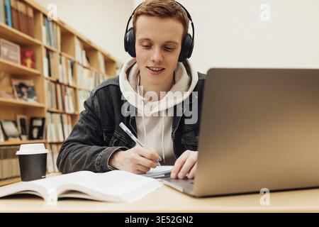 Smiling guy in headphones sitting at desk in library and writing Stock Photo