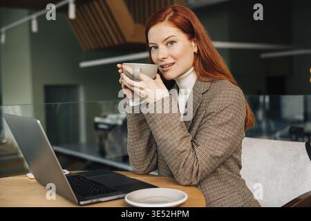 Young woman with ginger hair holding a cup. Female entrepreneur drinks coffee while working from a coffee shop Stock Photo