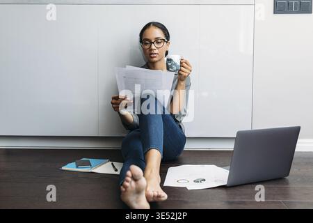 Woman looking on documents while drinking coffee at kitchen counter Stock Photo