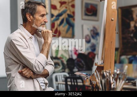 Side view of a thoughtful artist holding paintbrush while sitting in his studio. Male painter looking on canvas Stock Photo
