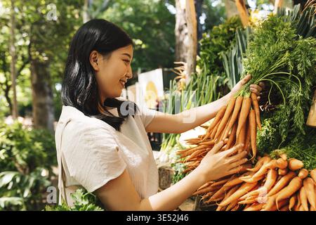 Smiling buyer holding a bunch of carrots. Asian woman choosing vegetables in the local market Stock Photo