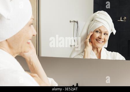 Aged woman with wrapped towel on her head touching cheek looking at a mirror Stock Photo