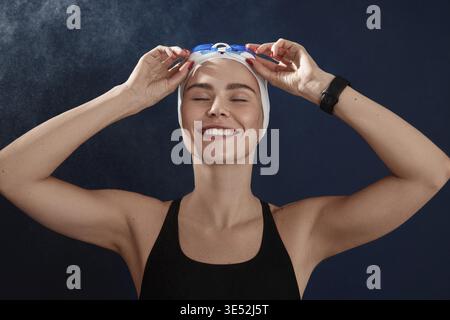 Young smiling female swimmer with closed eyes adjusting goggles standing over a blue backdrop Stock Photo