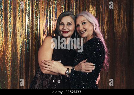 Two smiling senior girlfriends wearing evening dresses hugging each other while standing at glitter background Stock Photo