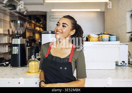 Beautiful smiling waitress standing at counter in coffee shop and looking away Stock Photo