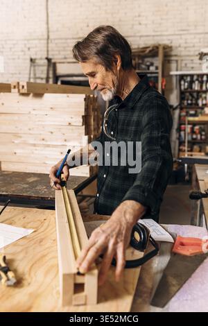 Side view of senior carpenter doing markings on a wooden bar with using a measuring tape Stock Photo