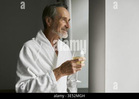 Smiling senior man with wineglass. Side view of a bearded mature male wearing a bathrobe in a hotel room looking at window Stock Photo