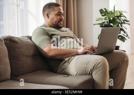 Smiling man with laptop on his hips sitting on a sofa. Young male working from home Stock Photo