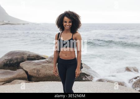 Smiling woman in fitness wear posing against the ocean after training Stock Photo