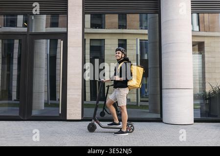 Smiling messneger man standing outdoors with electric push scooter. Delivery man with termal backpack in the city Stock Photo