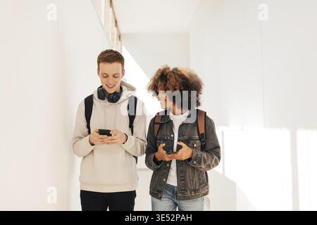 Two smiling students walking in corridor and chatting while holding smartphones Stock Photo