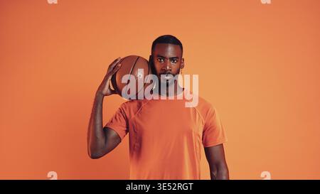 Serious basketball player looking at camera. Portrait of a male with basket ball on his shoulder against an orange background Stock Photo