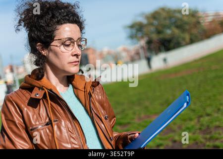 Professional businesswoman with glasses studies documents from a blue folder outdoors in a sunny urban park, focused and thoughtful in smart casual at Stock Photo