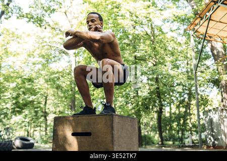Athlete doing box jumping on sports ground. Young male performing squats on wooden box Stock Photo