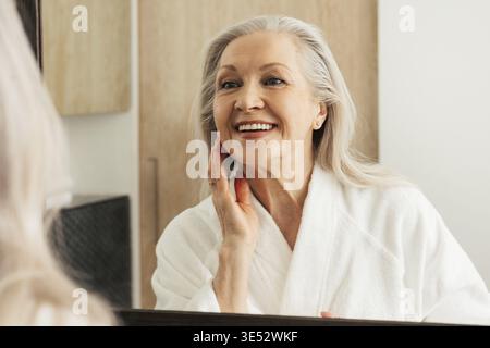 Happy aged female touching her face in front of a mirror. Woman in bathrobe admires her reflection Stock Photo
