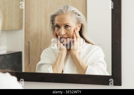 Senior woman in a bathrobe looking at a mirror and touching the face Stock Photo