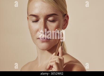 Young woman with closed eyes using face massager. Female with smooth freckled skin using face roller against a beige background Stock Photo