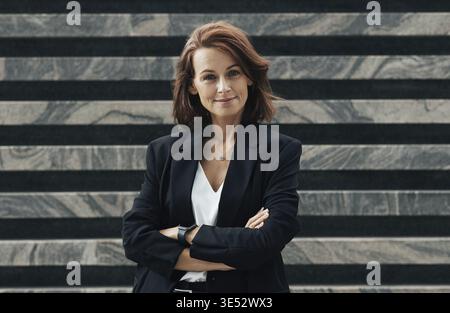 Smiling and confident middle-aged female with crossed hands. Businesswoman in black formal clothes with ginger hair Stock Photo