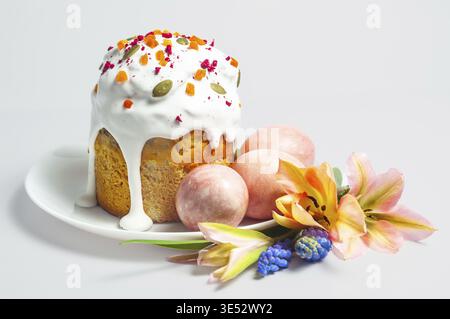 A festive Easter kulich adorned with icing and toppings, surrounded by pastel-colored eggs and delicate blooming flowers, presented on a clean white s Stock Photo
